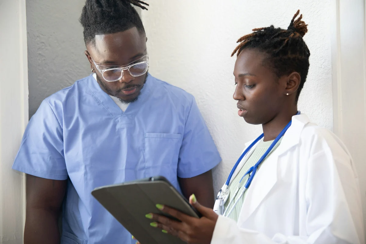 Two health care workers, one in blue scrubs and one in a white coat with a stethoscope, review information together on a tablet