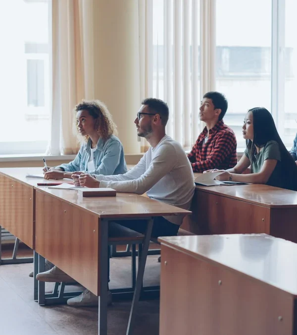 An instructor talks with a class of adult students taking notes at desks in a bright college classroom